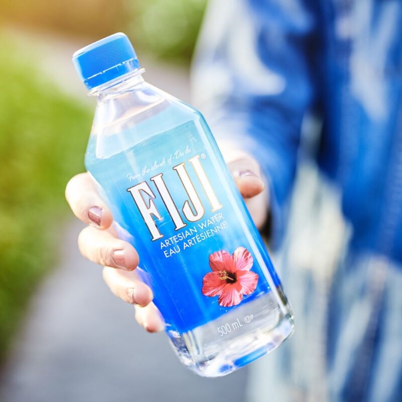 Kvinde holder en Fiji Vand i hånden, og rækker den frem mod dig.
